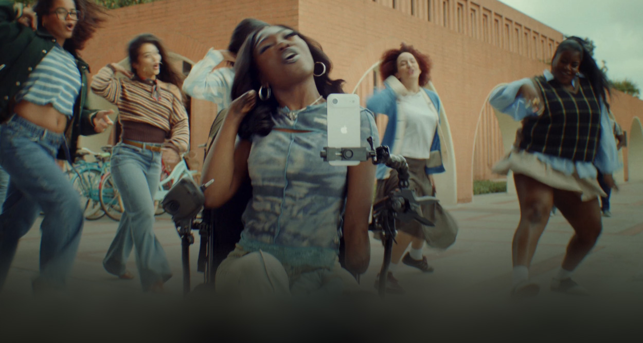 Student in wheelchair using accessibility feature on iPhone, surrounded by other  students who are dancing